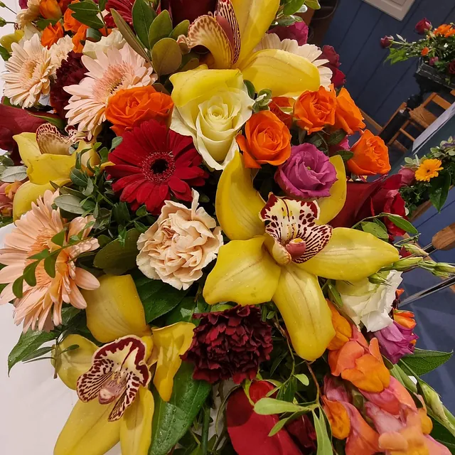 Stor blomsteroppsats med gule liljer, røde roser og oransje blomster i kirkemiljø.