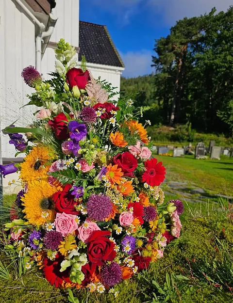 Fargerik blomsteroppsats med roser, solsikker og markblomster plassert ute ved kirke og gravplass.