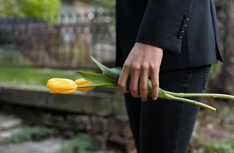 Person i mørk bekledning holder to gule tulipaner i hånden, i utendørs gravplassmiljø.
