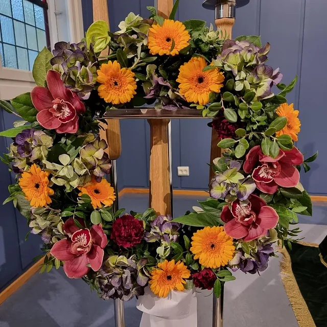 Rund krans med oransje, lilla og røde blomster dekorert med grønt, plassert inne i kirkerom.