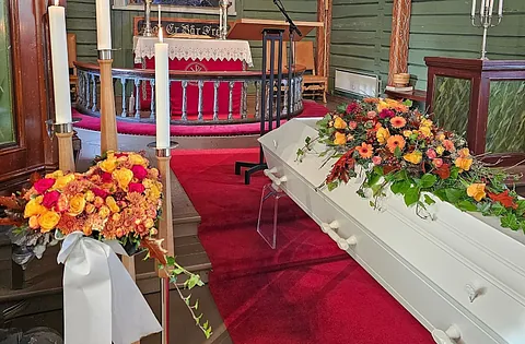 Hvit kiste med blomsterdekorasjon i en kirke med grønnmalte vegger, rødt teppe og tente lys.